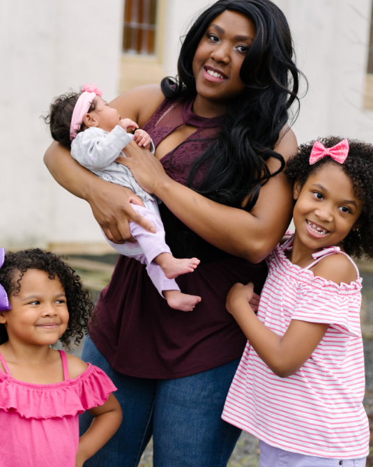 Afro latina mother holding infant with older daughter hugging side and younger daughter smiling