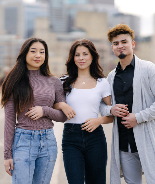 Two Latina young women and a Latino young man with link arms and cityscape in background