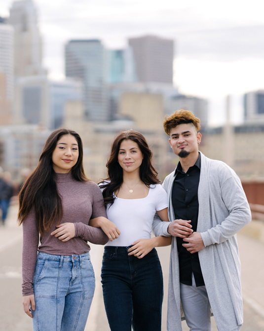 Two Latina young women and a Latino young man with link arms and cityscape in background