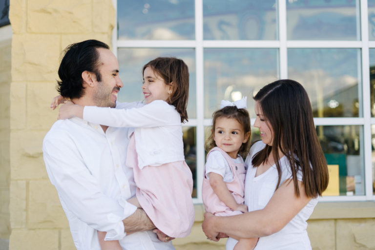 Father and mother holding and smiling two young daughters in front of a window